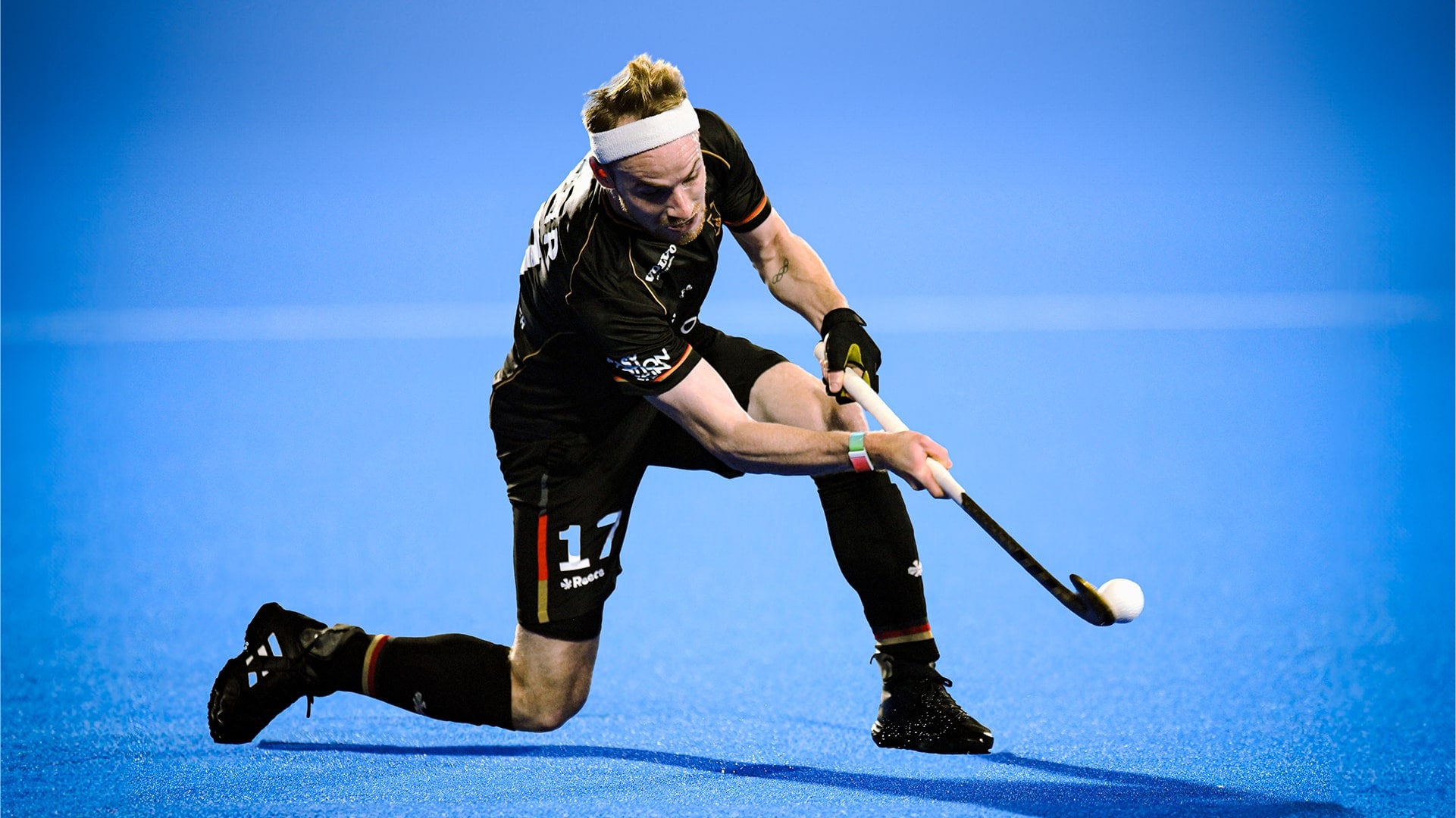 Christopher Rühr setzt auf einem blauen Hockeyfeld kniend zum Schlag an, wobei der Ball sich gerade an der Spitze seines Schlägers befindet.				 				 				 				 				 				 				 				 				 				 				 				